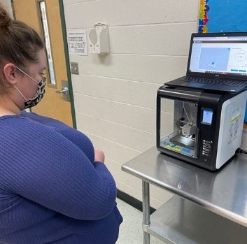 A woman stands to the left inside a classroom wearing a leopard print medical face mask and watches a 3D printer work.