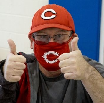 A man with glasses wearing a Cincinnati Reds hat and cloth face mask gives the camera a double-thumbs up.
