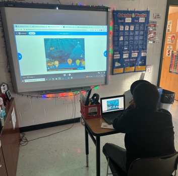 A student wearing a hat sits at a desk in a dark room with a laptop on his desk and looks up at a smartboard