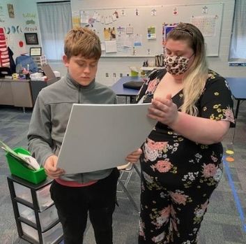A boy looks down at a large canvas while his teacher stands next to him. They are in a classroom.