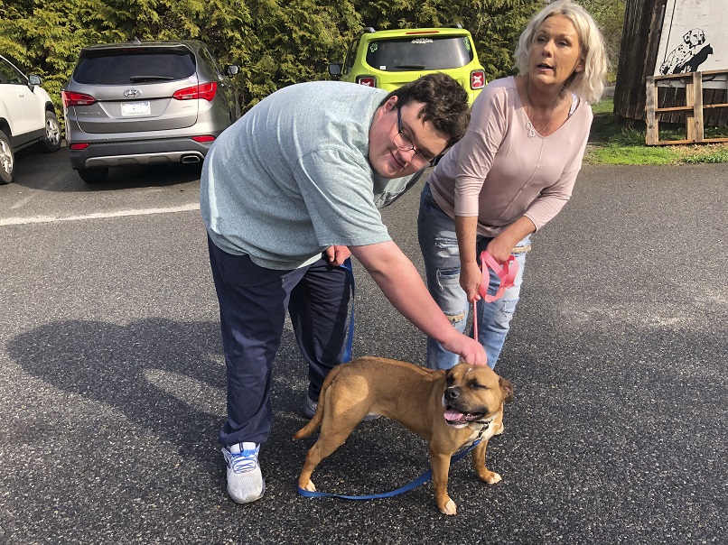 A teenage boy wearing glasses leans over and pets a dog on the head while a woman holds the leash.