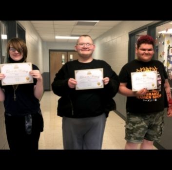 A young girl and two young boys stand in a line in a school hallway smiling and each holding up a certificate