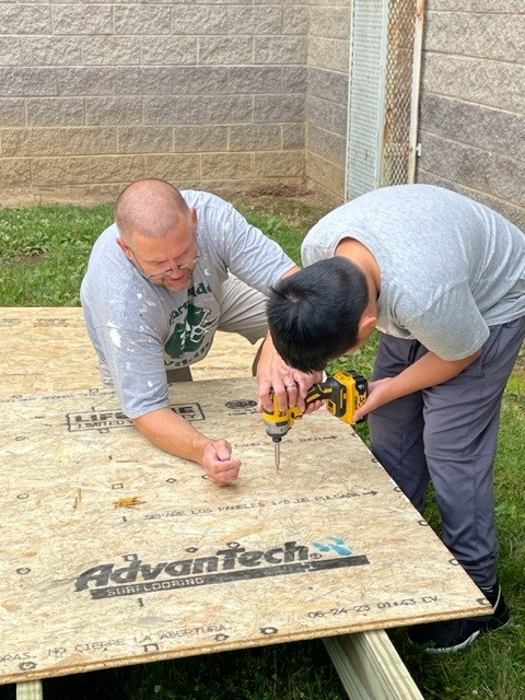 A man and a teen boy work together to drill into a piece of plywood