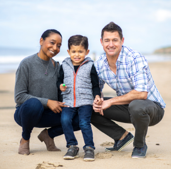 An African American woman and Latino man pose on the beach with a Latino toddler. They are dressed for autumn.