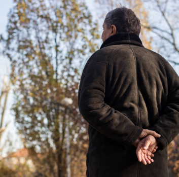 A man wearing a dark grey coat stands with his back to the camera. His hands are clasped behind is back and he is looking at trees in the distance.