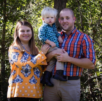 A man holding a toddler stands next to his wife outside. They are all smiling at the camera.