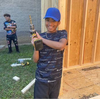 A black tween wearing a blue baseball cap smiles and points a drill into the air