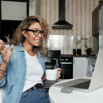 A woman wearing glasses and holding a mug participates in a video chat from her kitchen table