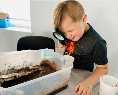 A child squints into a magnify glass looking into a plastic container of dirt and rocks