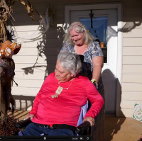 A woman wearing a pink shirt is pushed in a wheel chair by another smiling woman