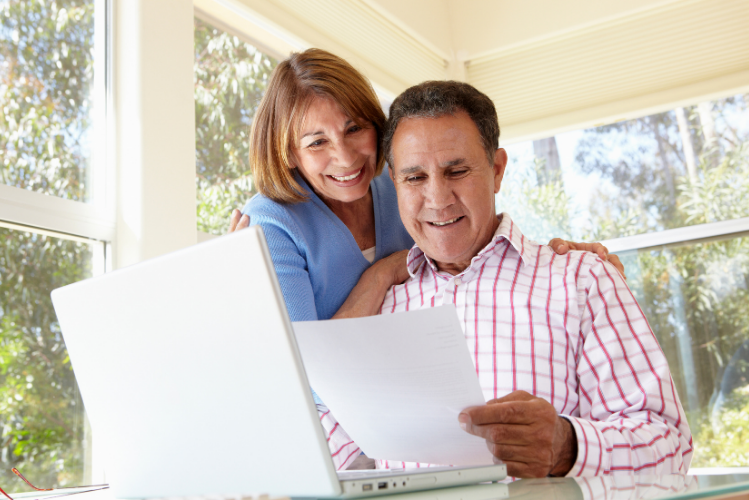 A woman looks over the shoulder of a man who is looking at a paper. They are in a sunroom and there is a laptop on the table in front of them.